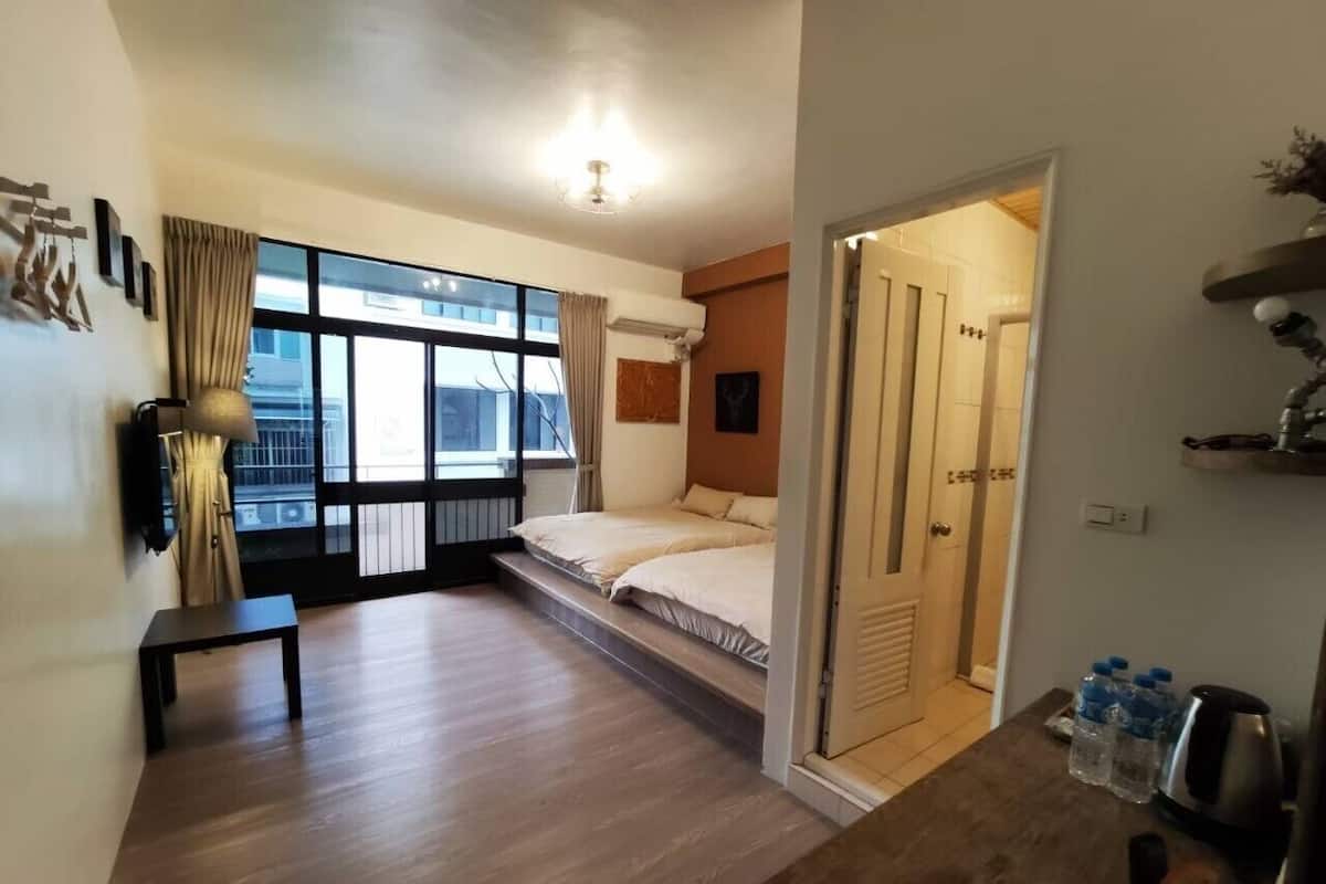 A modern bedroom with a large window, a bed, a small table, and a kitchenette area.