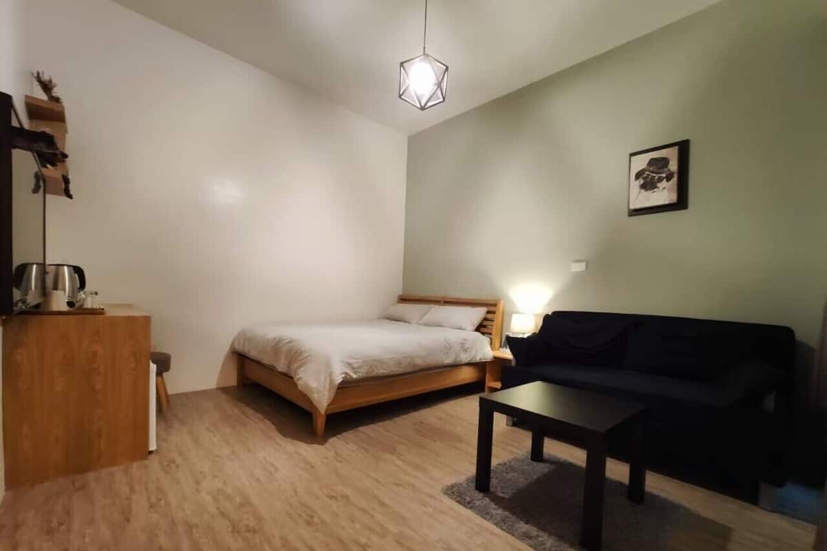 A compact room with a bed, a wooden desk, a black sofa, and a hanging light fixture.