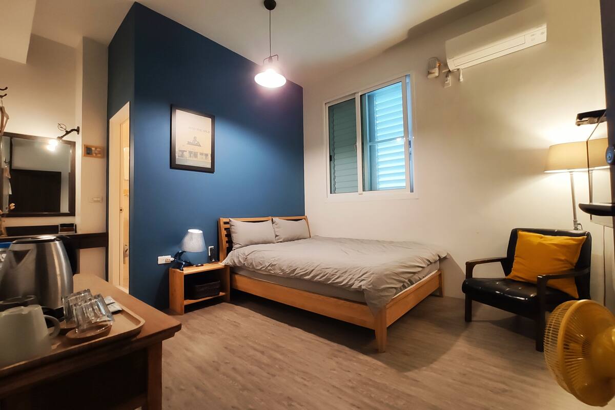A bedroom with a bed, bedside table, lamp, chair, and a fan.