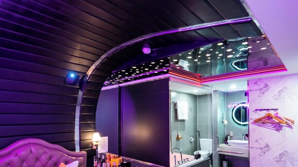 A modern bedroom with a curved ceiling, a large bed with pillows, a nightstand, and a bathroom with a glass shower enclosure. - Cupid Hotel (Ho Chi Minh City)