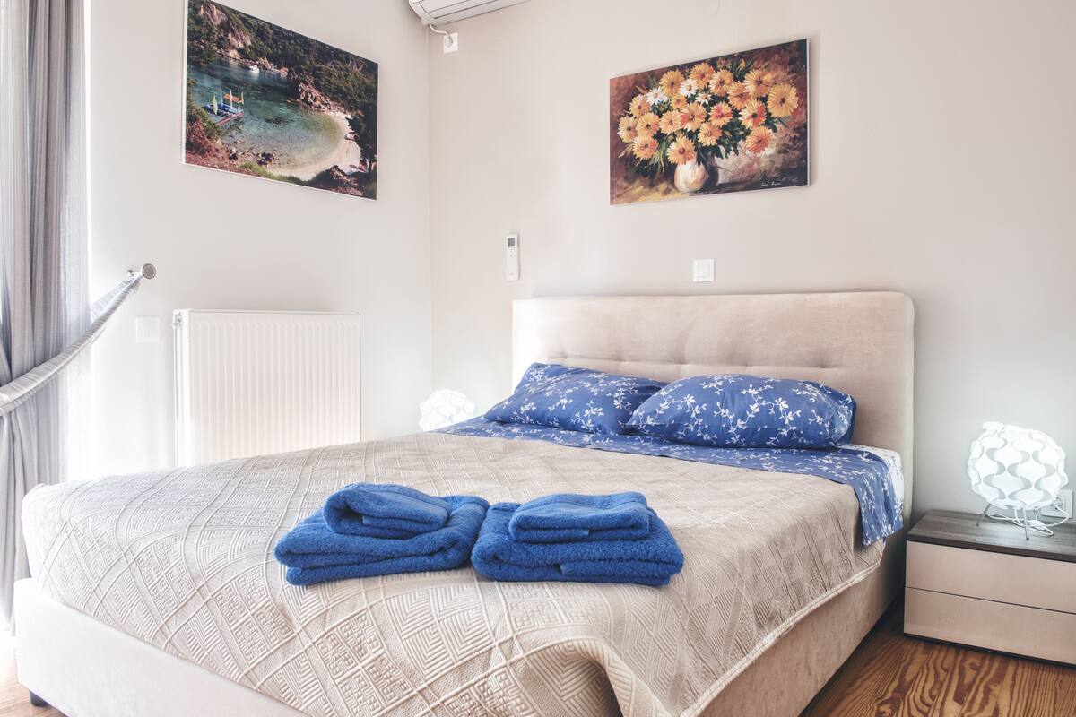 A neatly made bed with a patterned bedspread and blue pillows, a pair of folded towels on the bed, and two wall-mounted paintings depicting a coastal scene and a bouquet of flowers.