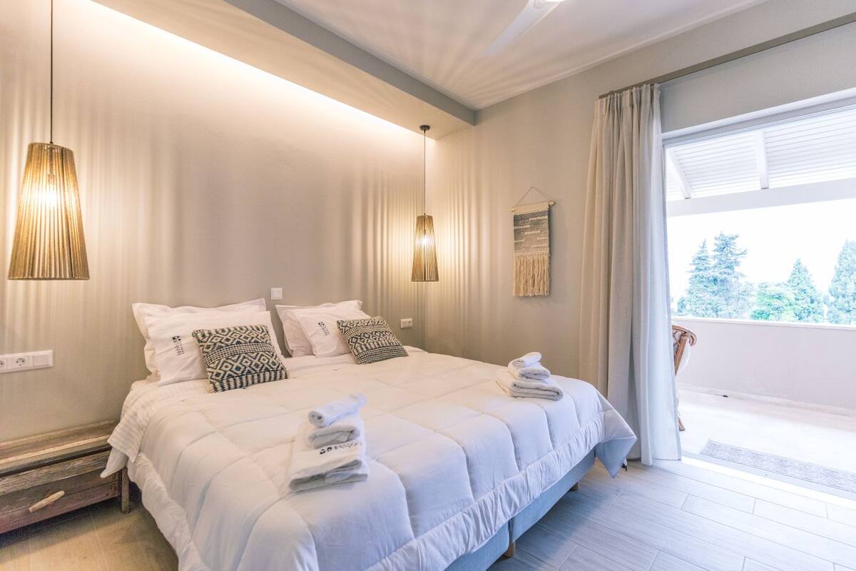A modern bedroom with a large bed, bedside lamps, a view of the outdoors, and a balcony.