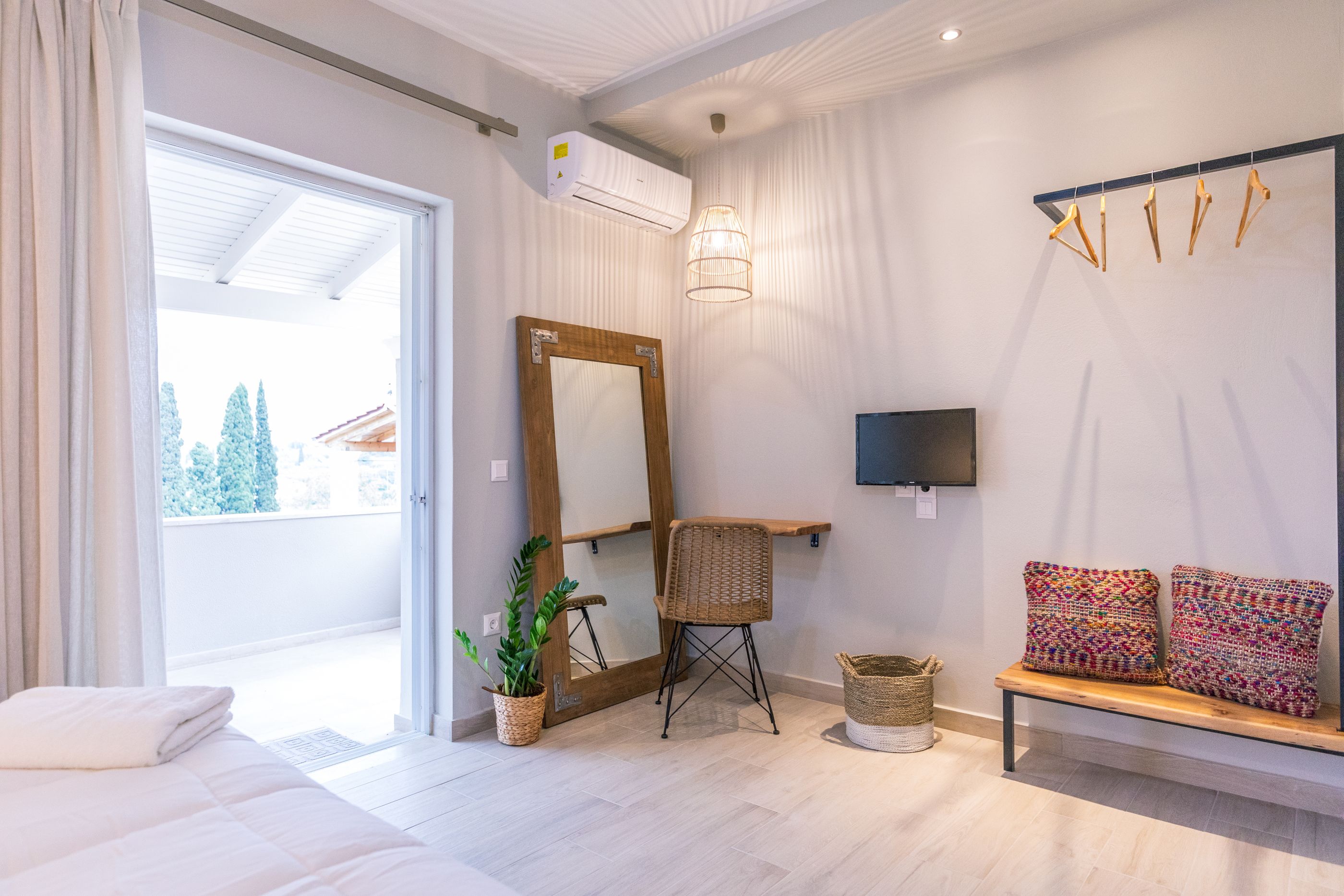 Ein modernes Hotelzimmer mit einem großen Spiegel, einer Holzbank mit bunten Kissen, einer Topfpflanze und einem an der Wand montierten Fernseher.