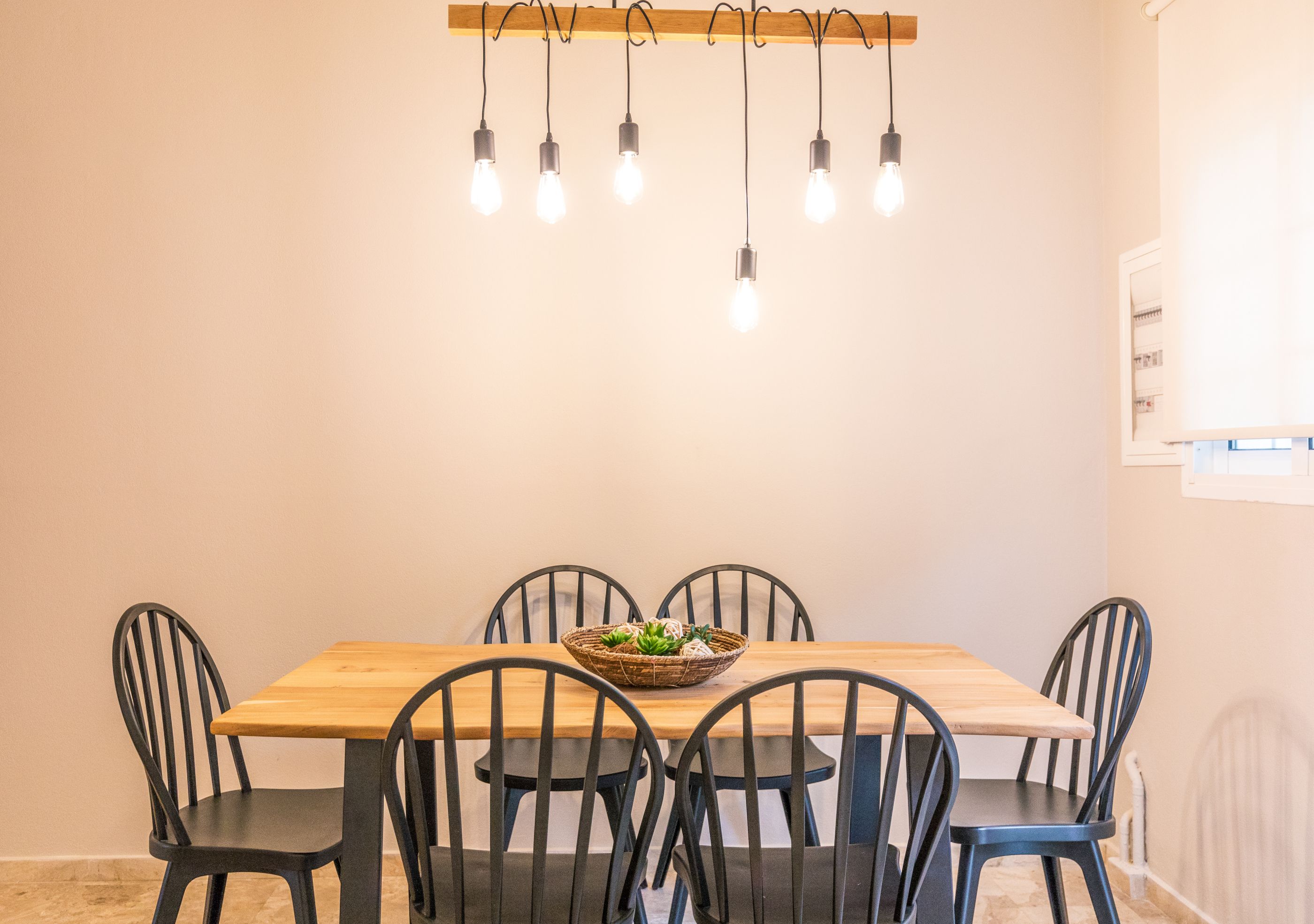 A dining area with a wooden table, six chairs, a basket of vegetables, and a row of hanging light bulbs.