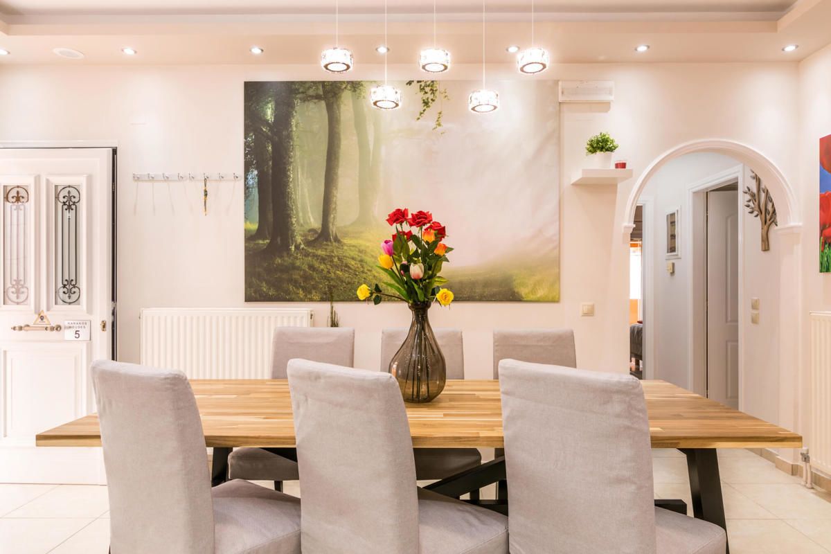 A dining area with a wooden table, six grey upholstered chairs, a vase with flowers, and a forest-themed painting on the wall.