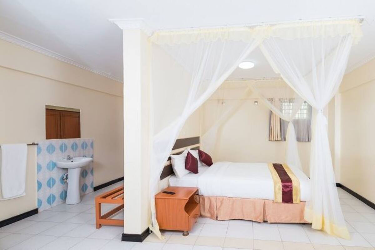 A hotel room with a bed under a canopy, a bedside table, a small wooden stool, a sink, and a mirror.