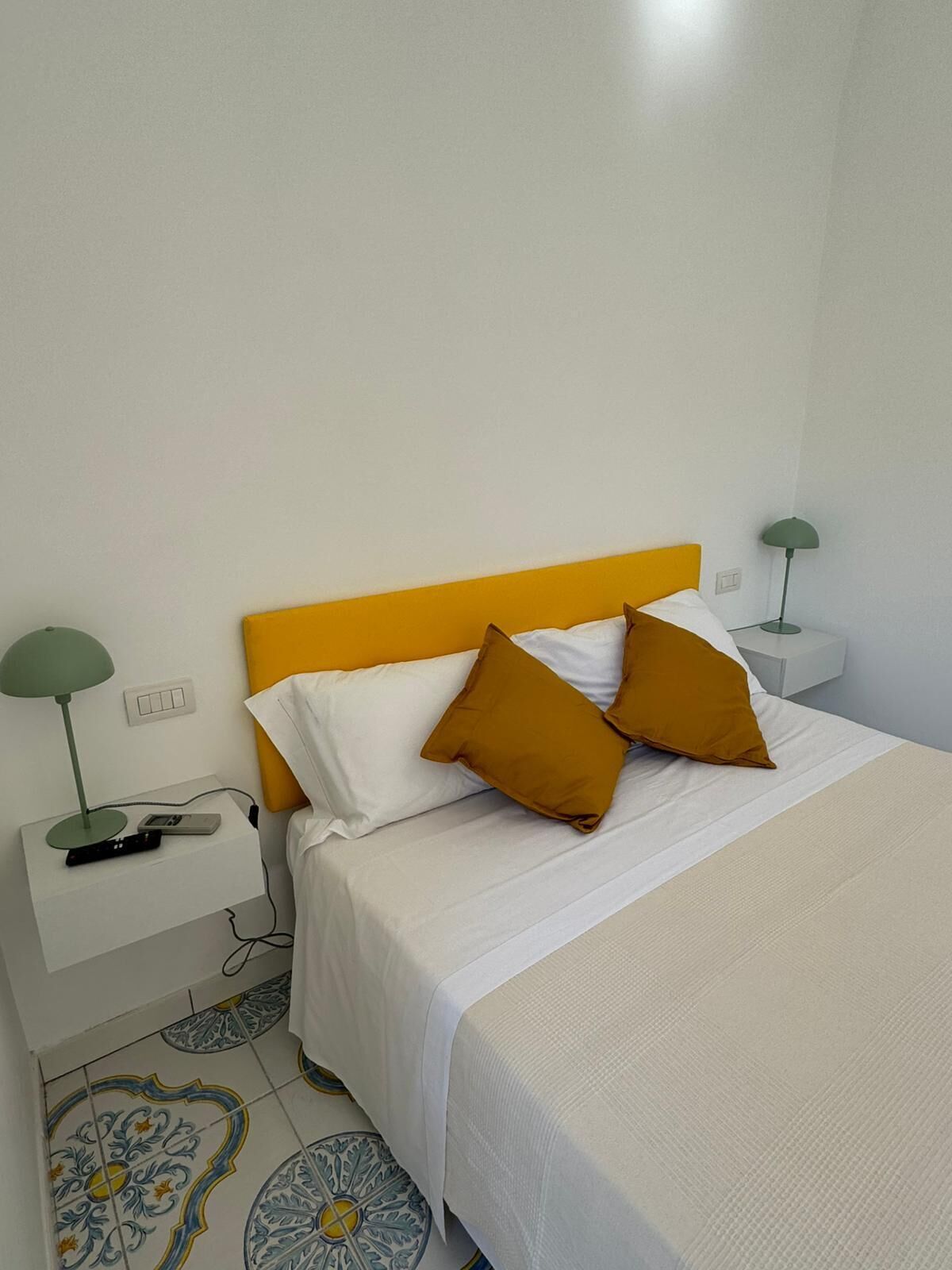 Una cama bien hecha con ropa blanca y dos almohadas color mostaza, una mesita de noche con una lámpara e un enchufe.