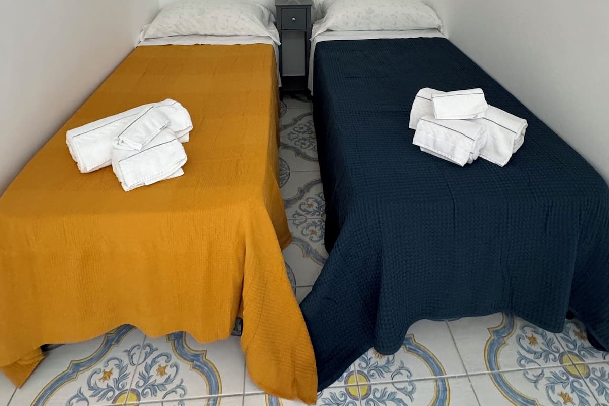 Ein Zimmer mit zwei Einzelbetten, jeweils ausgestattet mit einem ordentlich gefalteten Handtuch und einem Kopfkissen. Dazwischen steht ein Nachttisch mit einer Lampe. Der Boden ist mit Fliesen in einem dekorativen Muster versehen.