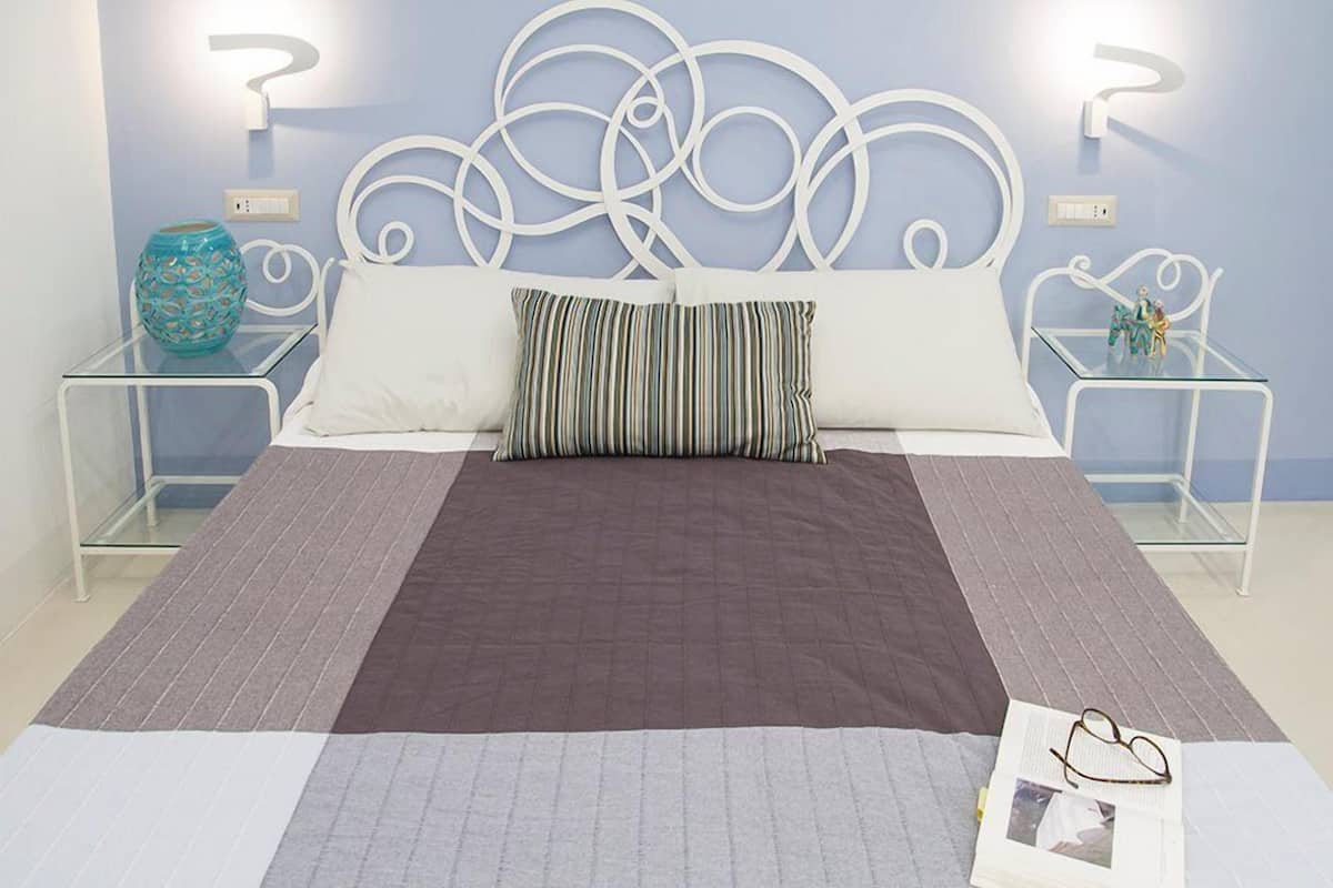 Ein ordentlich bezogenes Bett mit einem braun-weiß gestreiften Kopfkissen, ein Glastische mit einem dekorativen Blumenvasen und eine Wand mit abstrakter Metallkunst.