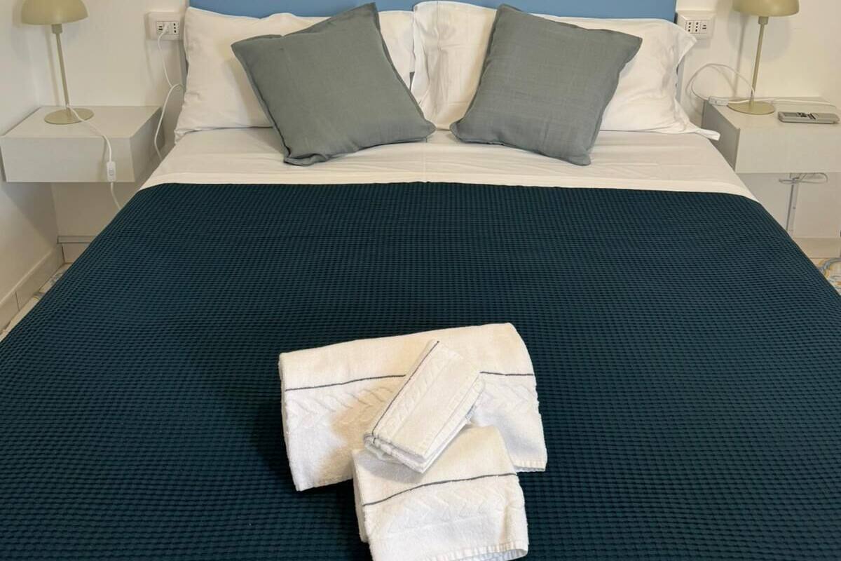 Ein ordentlich bezogenes Bett mit blauem Kopfteil, zwei grauen Kissen und einem gefalteten weißen Handtuch auf einer dunkelblauen Bettdecke.