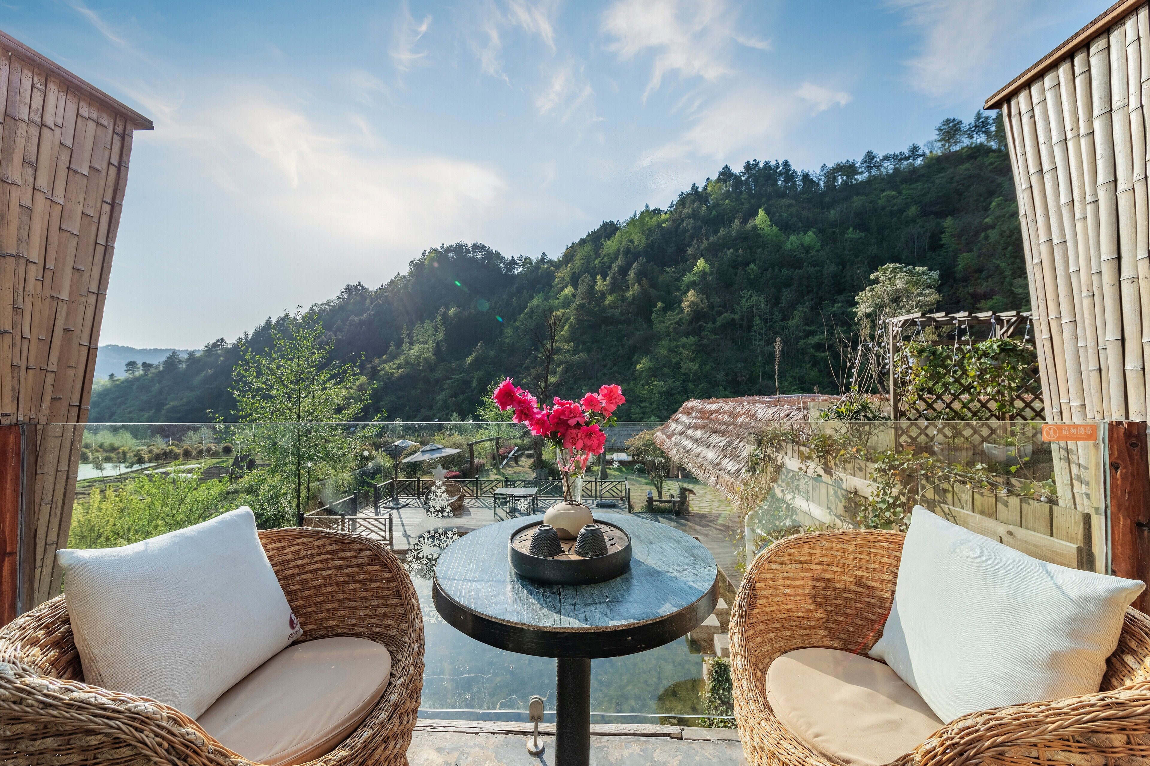 A balcony with wicker chairs and a small table, overlooking a lush green landscape and a river.