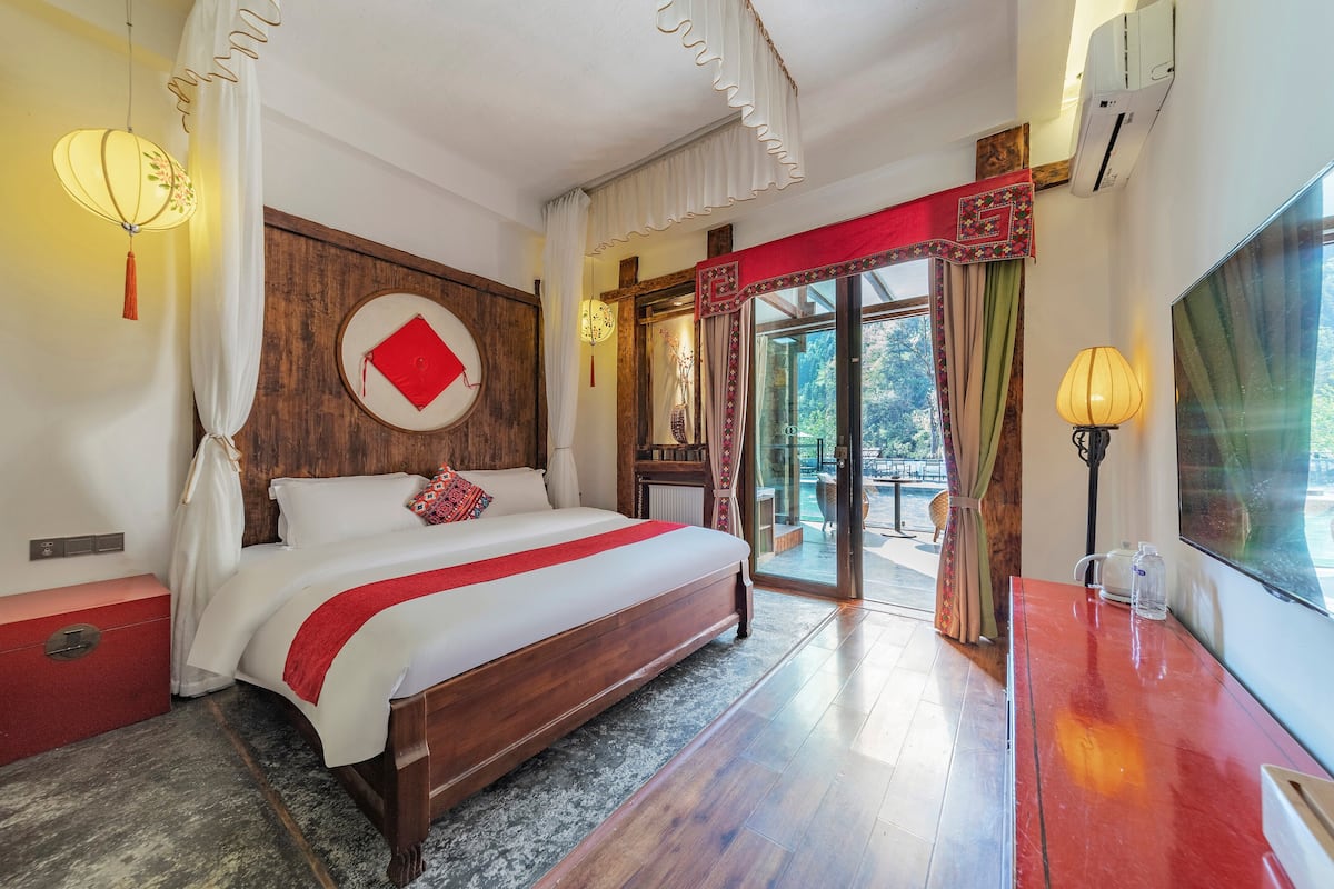 Habitació d'hotel amb un llit gran, un capçal de fusta, un coixí decoratiu vermell, una tauleta de nit vermella, aire condicionat fixat a la paret i vista a l'exterior a través d'una porta de vidre.