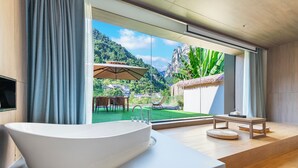 A modern bathroom with a large bathtub, a wooden vanity, and a view of a lush garden and mountains through large windows. - Pandora Vista Retreat  (Zhangjiajie)