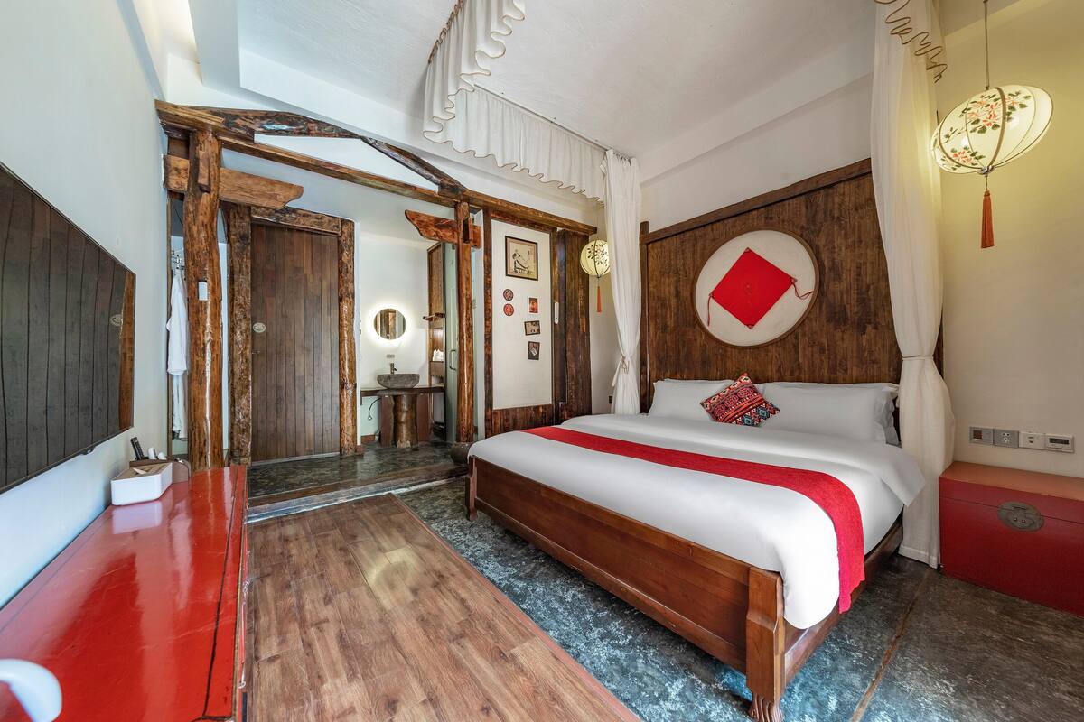 Une chambre avec un lit en bois, un coussin rouge, une couvre-lit blanche et une couverture rouge. On y trouve une commode en bois, une chaise rouge et une petite table avec une lampe. La pièce a un plancher en bois et un plafond blanc avec un luminaire.