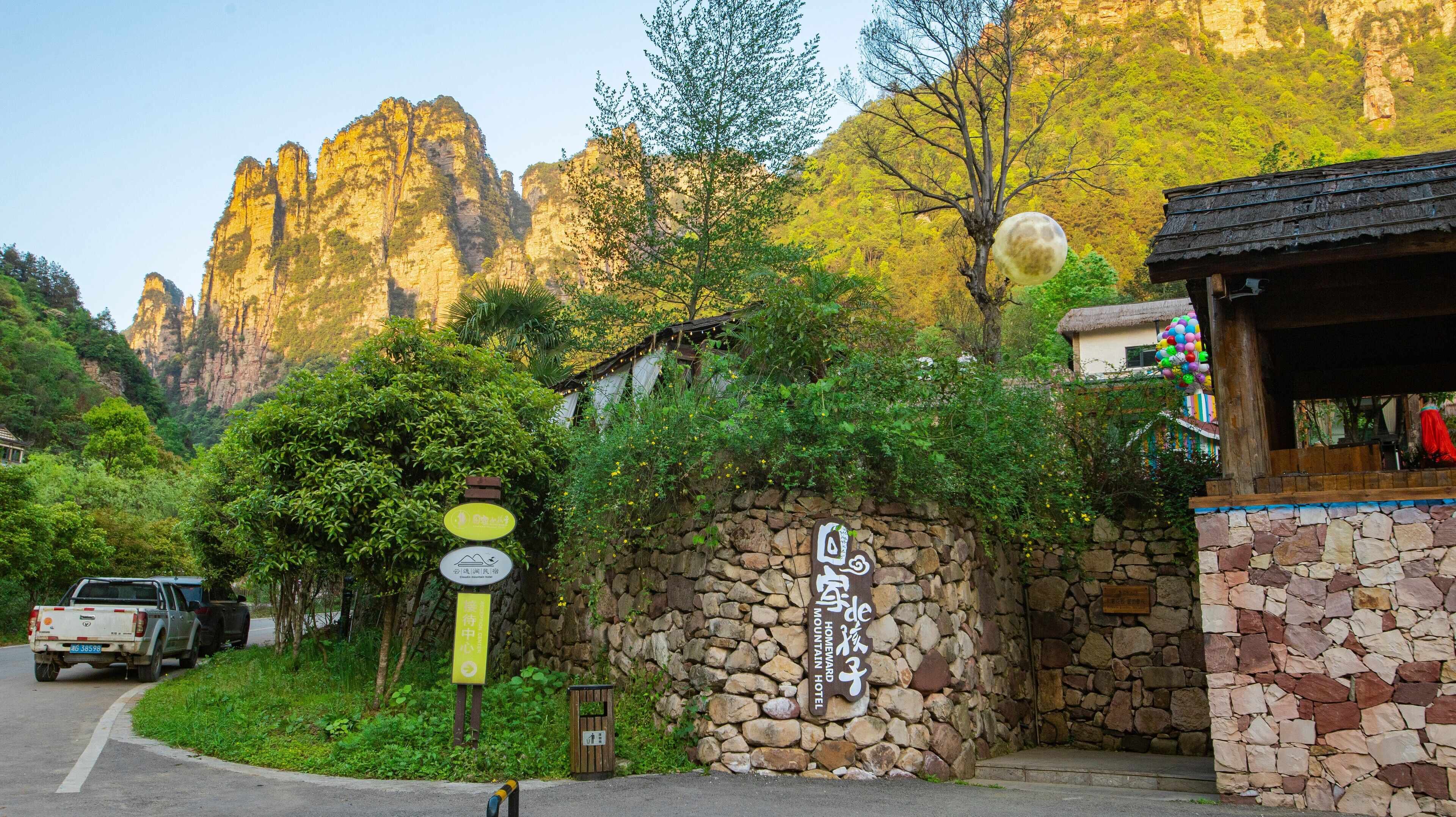 A stone wall with a sign for a mountain hotel, a thatched-roof building, and a road with a truck.
