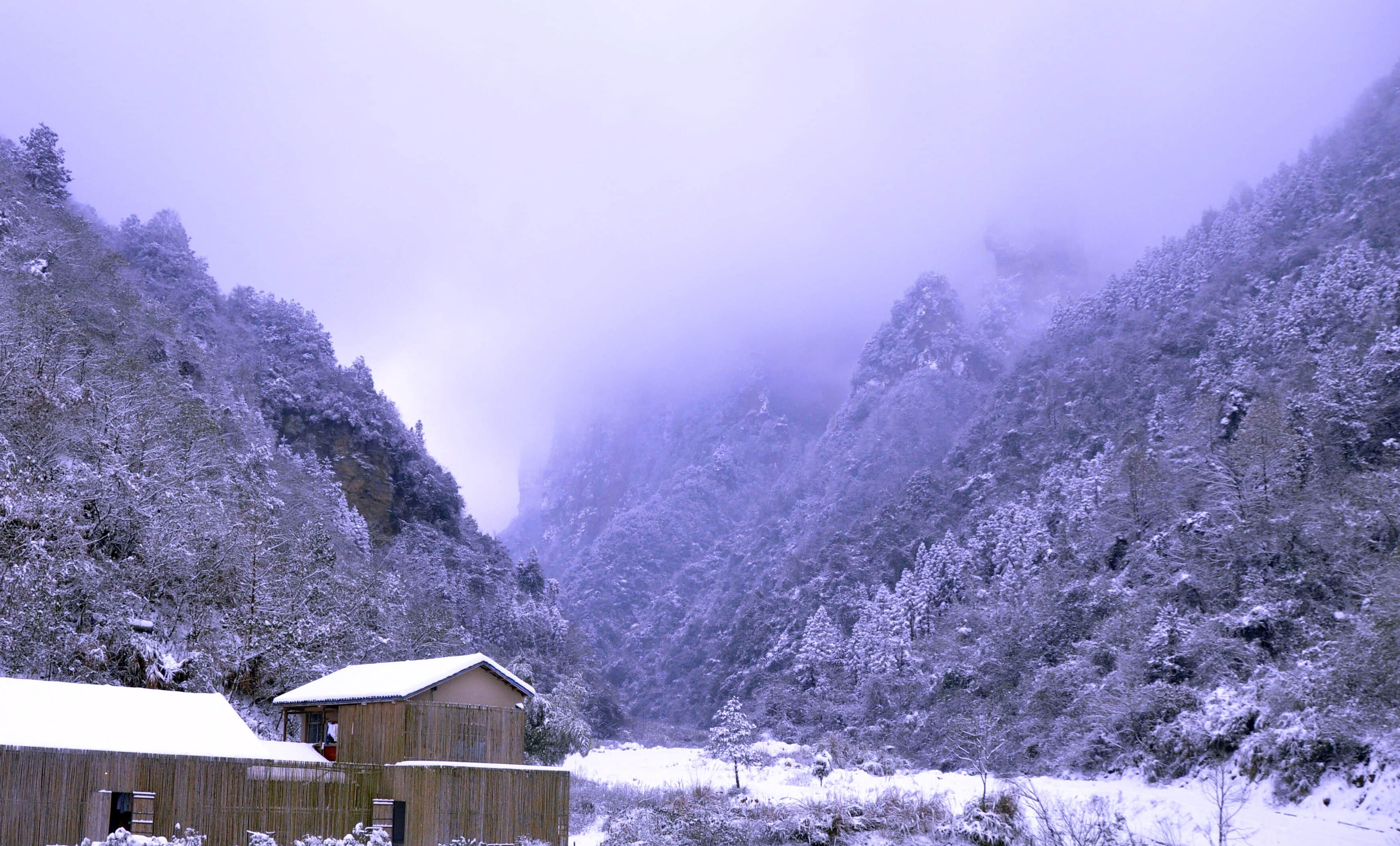 A wooden cabin with a snow-covered roof, surrounded by snow-covered trees and a mountainous backdrop.