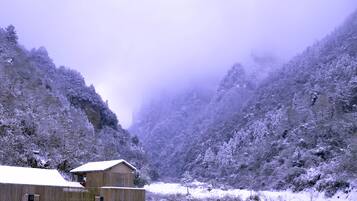 A wooden cabin with a snow-covered roof, surrounded by snow-covered trees and a mountainous backdrop.