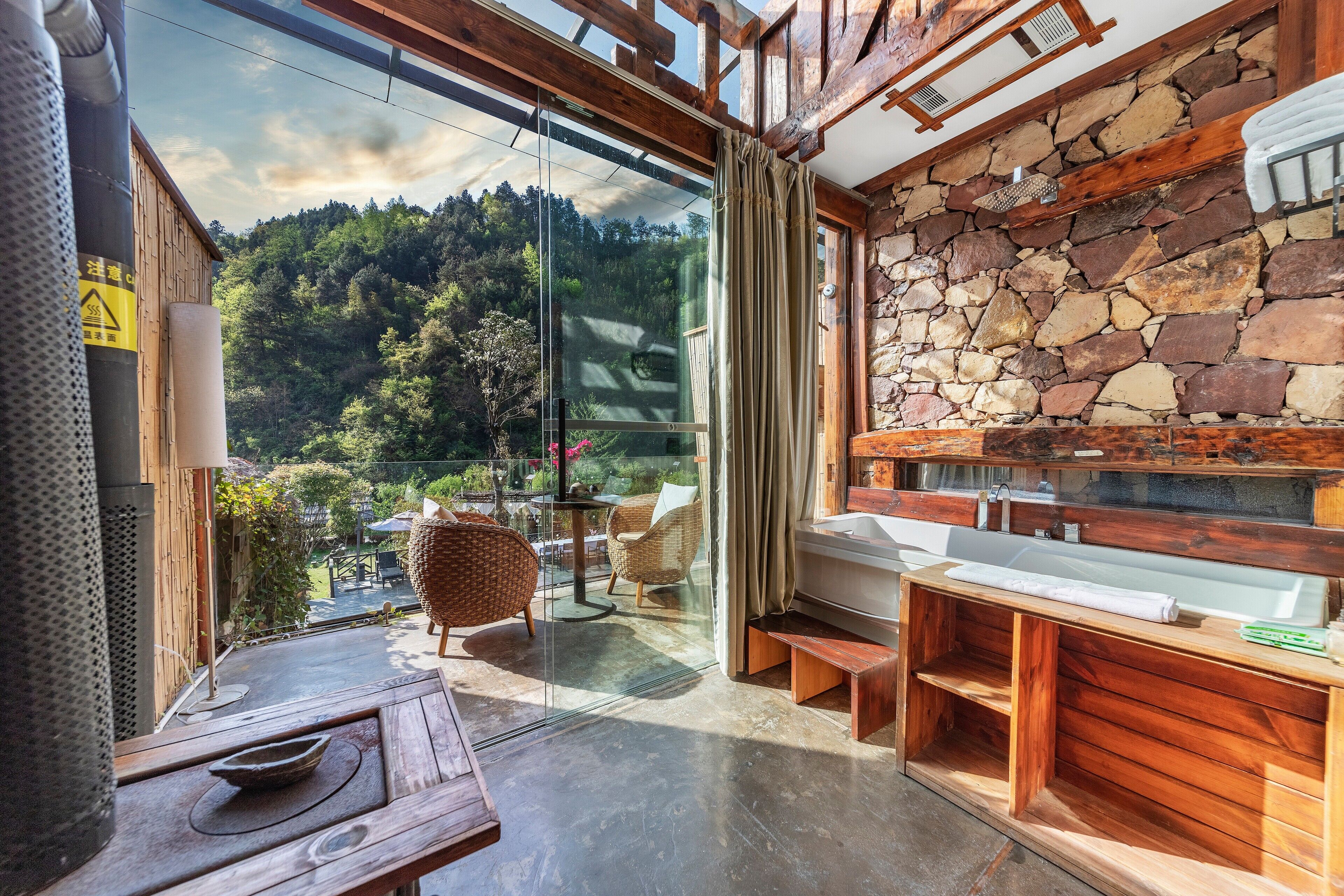 Ein modernes Badezimmer mit Steinwand, Holztischbein und großem Fenster mit Blick auf ein bewaldetes Gebiet.