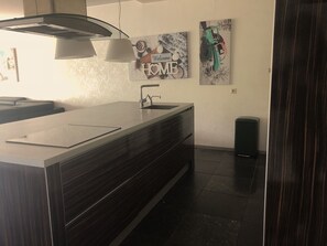 Cozinha moderna com armário de madeira escura, pia de aço inoxidável e bancada branca. Possui duas luminárias fixadas na parede e duas fotos emolduradas.