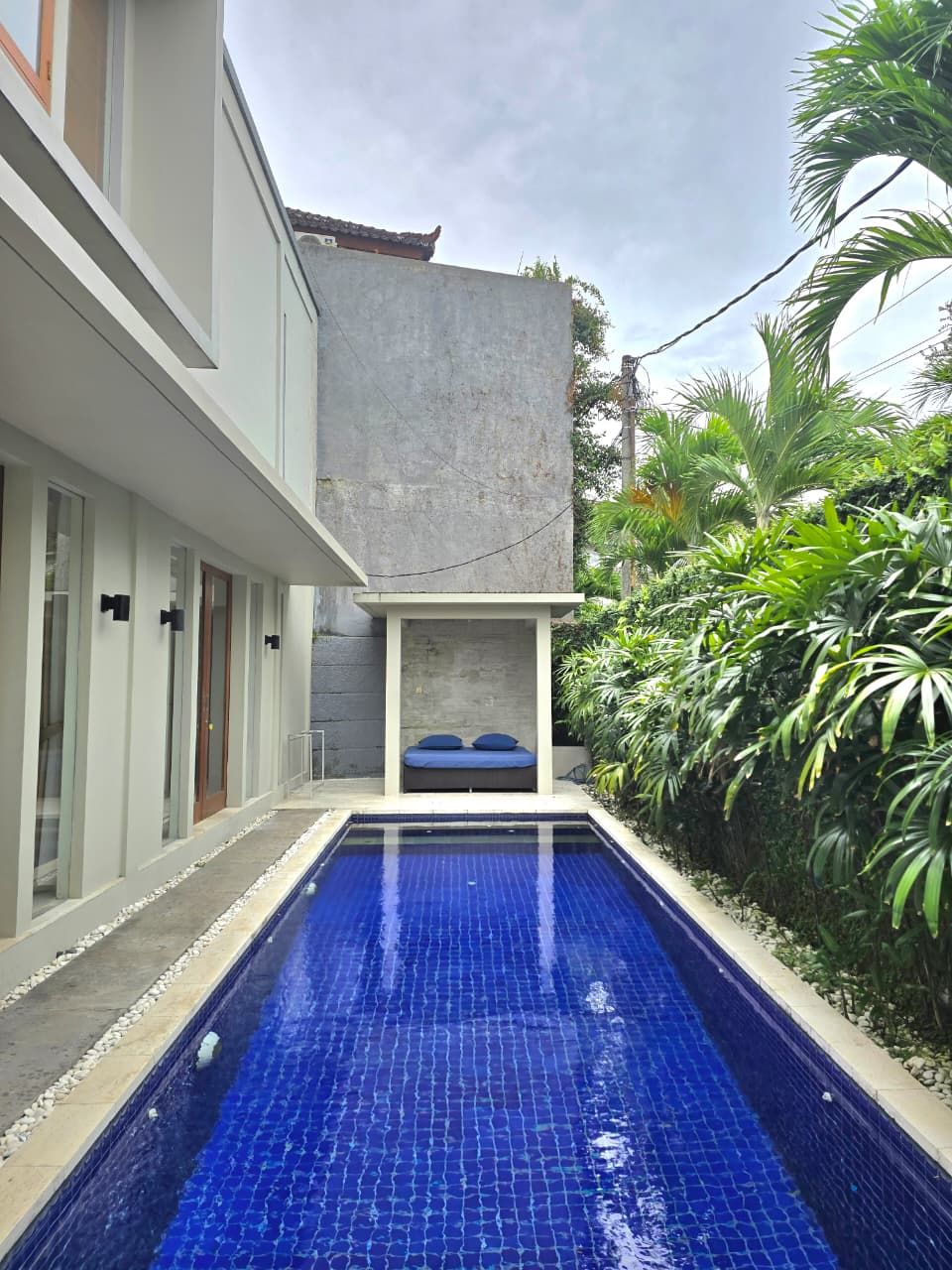 A modern building with a swimming pool, a small seating area with cushions, and lush greenery around.