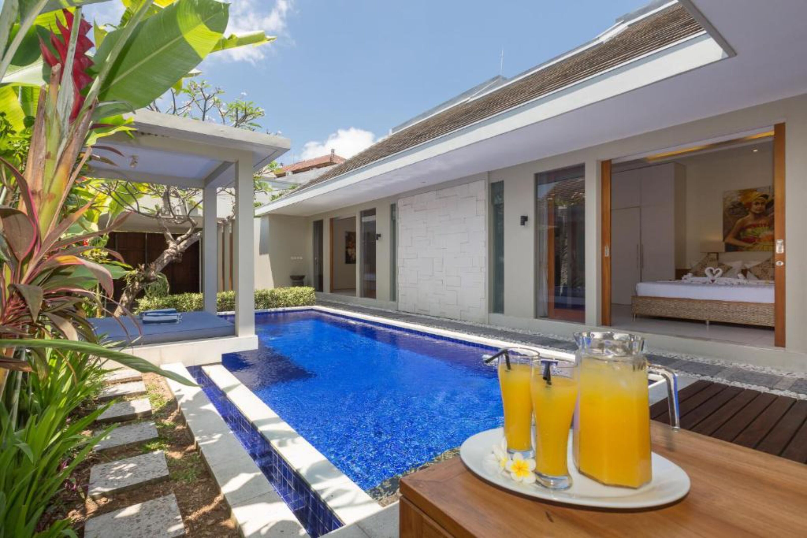 A modern poolside area with a clear blue pool, a wooden table with drinks, and a white exterior wall.