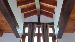 Ein Zimmer mit schräg verlaufender Holzbalkendecke, freiliegenden Balken und großen Fenstern mit Vorhängen.