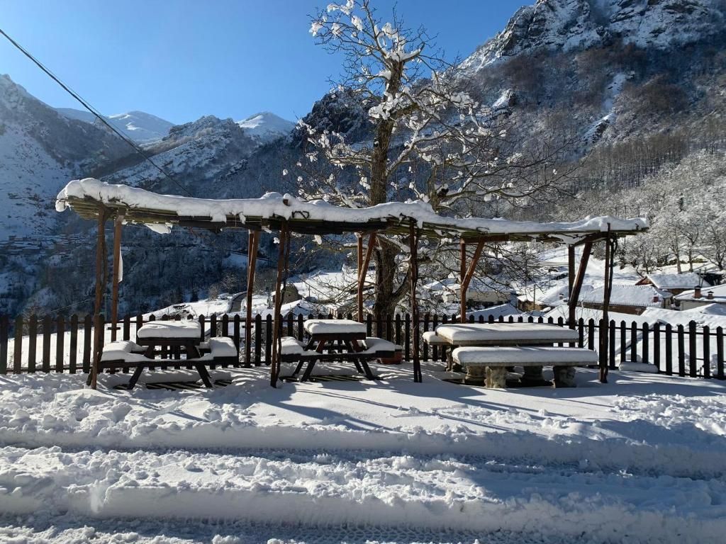 Un espacio al aire libre cubierto de nieve con mesas de picnic y bancos, rodeado por una valla de madera, con montañas al fondo.