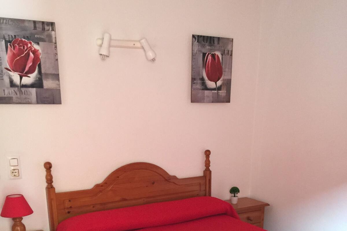 Un dormitorio con una cama de madera, una colcha roja, una mesita de noche con lámpara y dos cuadros colgados en la pared.