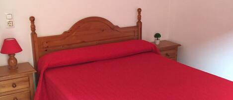 Un dormitorio con una cama de madera, una colcha roja, una mesita de noche con lámpara y dos cuadros colgados en la pared.