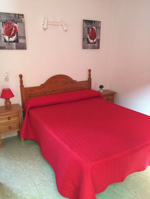 Une chambre à coucher avec un lit en bois, une couverture rouge, une table de chevet avec une lampe et deux tableaux accrochés au mur.