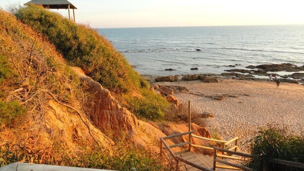 A beach with a wooden staircase leading down to the shore, a gazebo on a grassy hill, and rocky outcrops in the water. - Hostal La Campa (Chiclana de la Frontera)