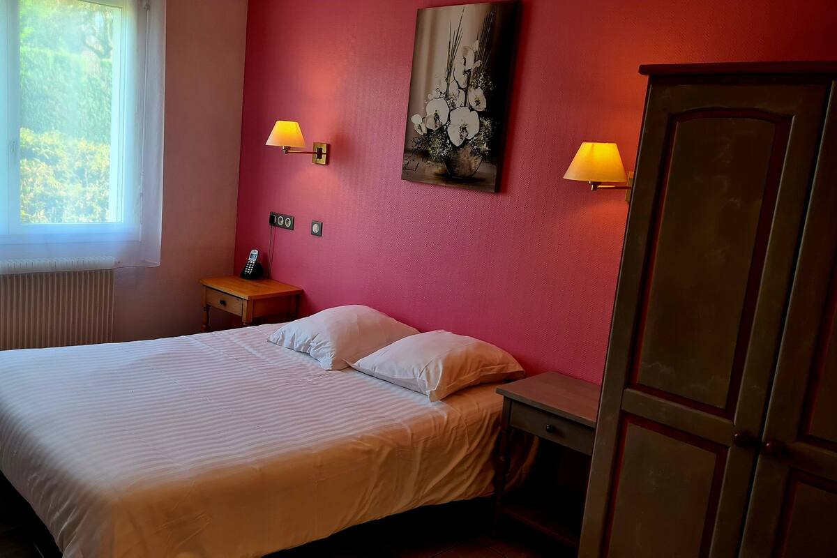 Een slaapkamer met een bed, twee bedlampen, een nachtkastje, een radiator en een raam met uitzicht op groen.