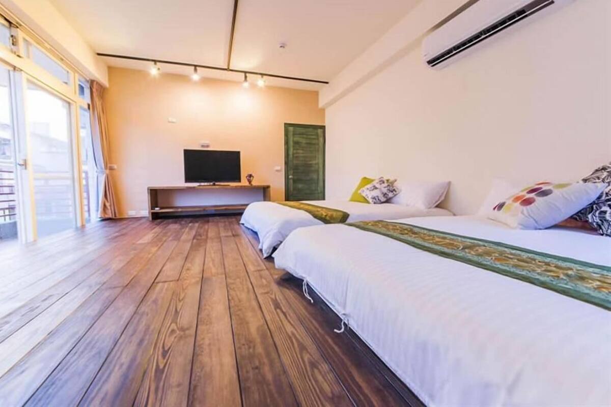 A hotel room with a wooden floor, two beds, a TV, and a sliding glass door to a balcony.