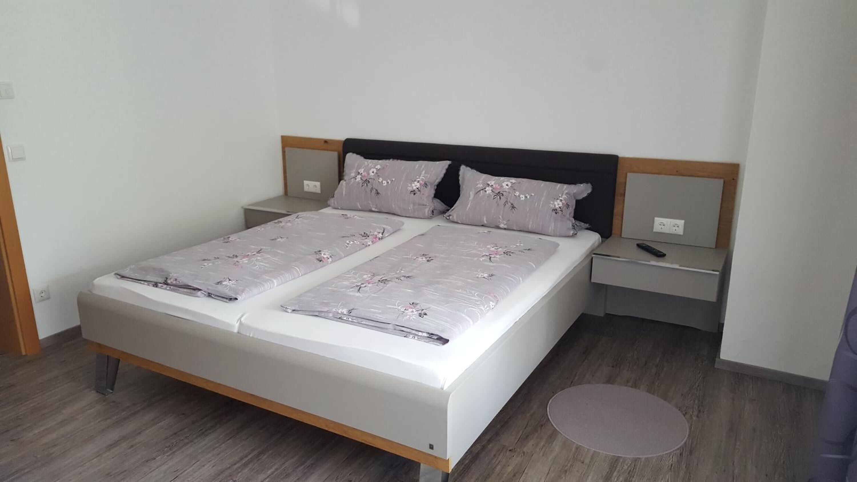 Een tweepersoonsbed met een houten hoofdbord, twee nachtkastjes en een klein rond vloerkleed.
