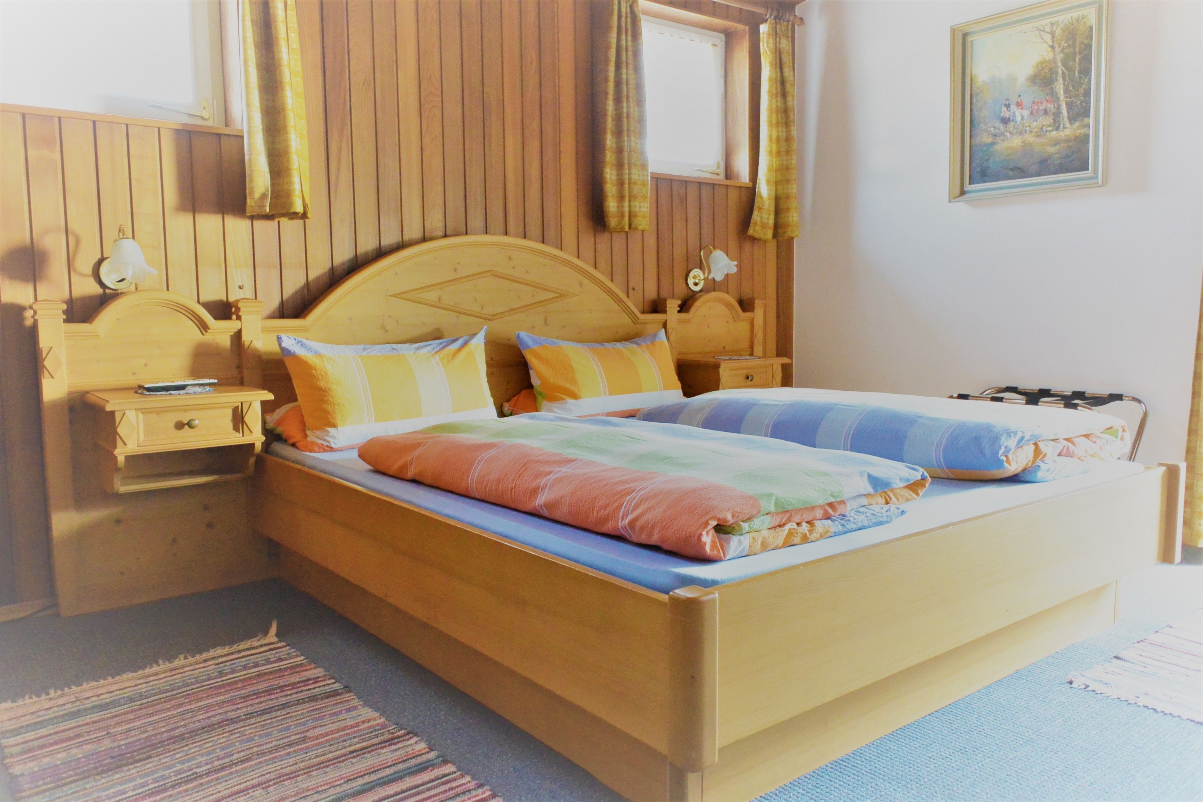 Una cama de madera con una colcha y almohadas de colores, una mesita de noche con una lámpara y una ventana con cortinas.
