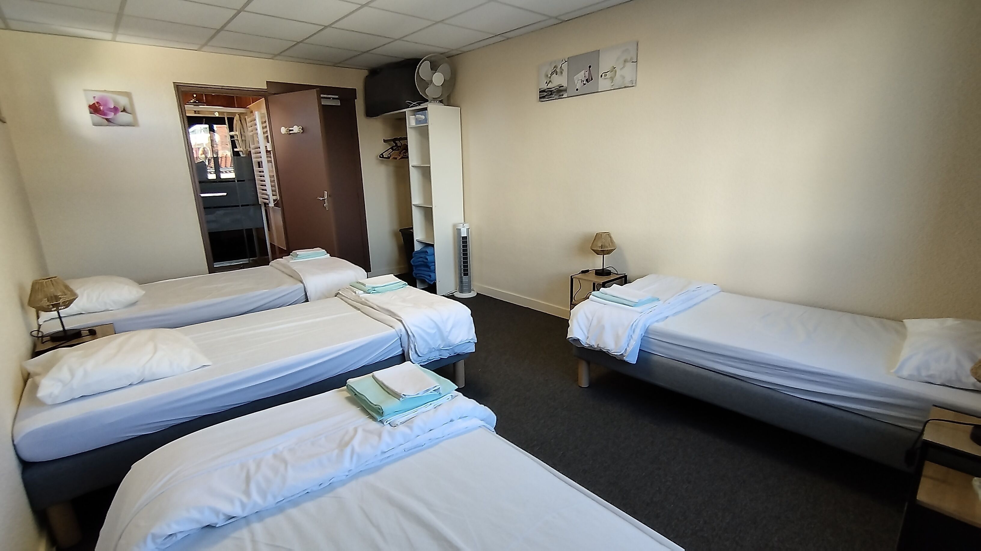 Habitación de hotel con tres camas individuales, un armario, un ventilador y una mesita de noche con lámpara.