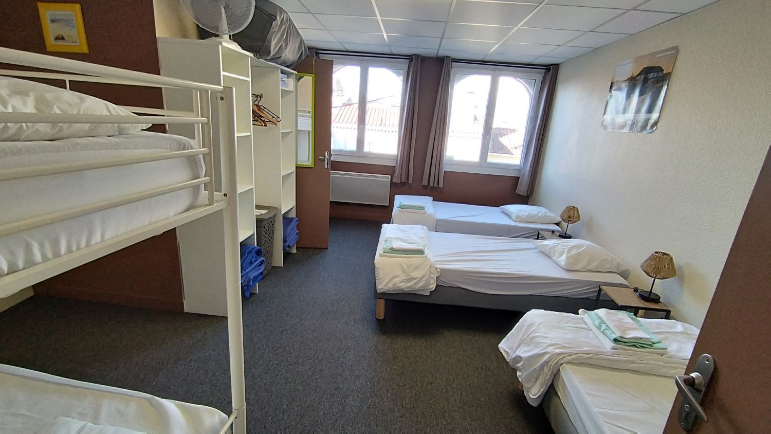 A hotel room with bunk beds, a wardrobe, a window with curtains, and a bedside table with a lamp.