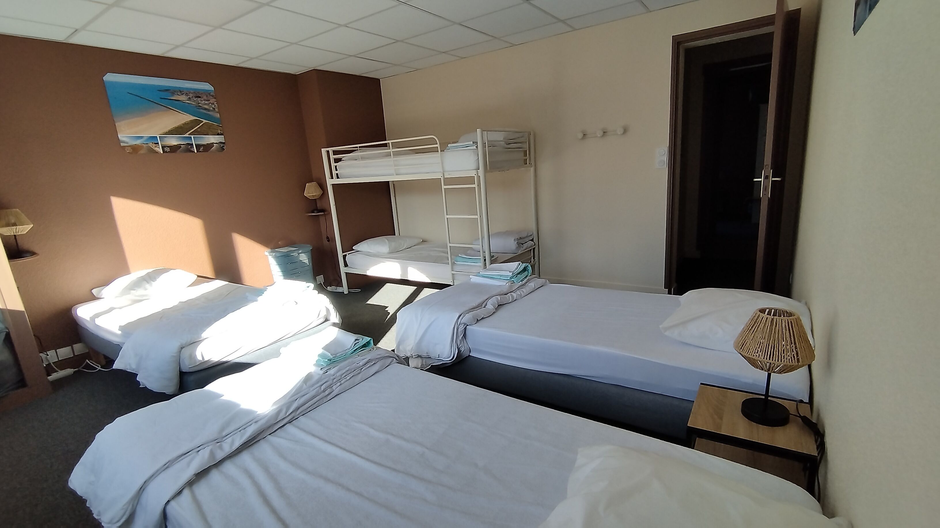 Une chambre d’hôtel avec deux lits simples, un lit superposé, un téléviseur fixé au mur, une porte et une petite table avec une lampe.