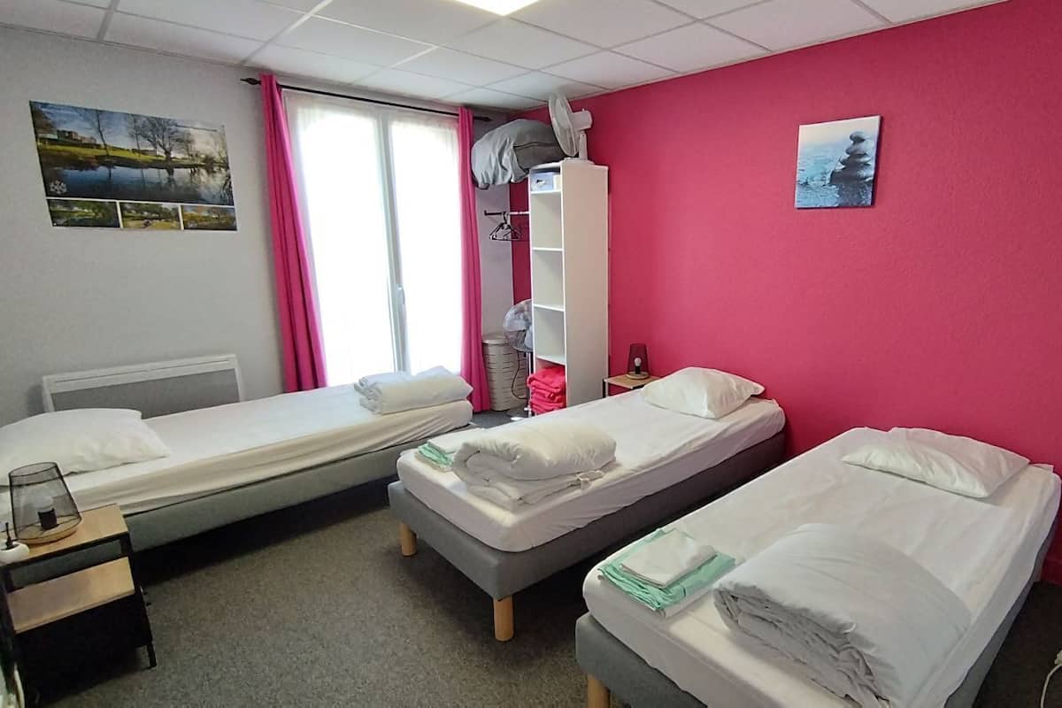 A room with three single beds, a window with pink curtains, a white fan, and a small shelf with a picture.