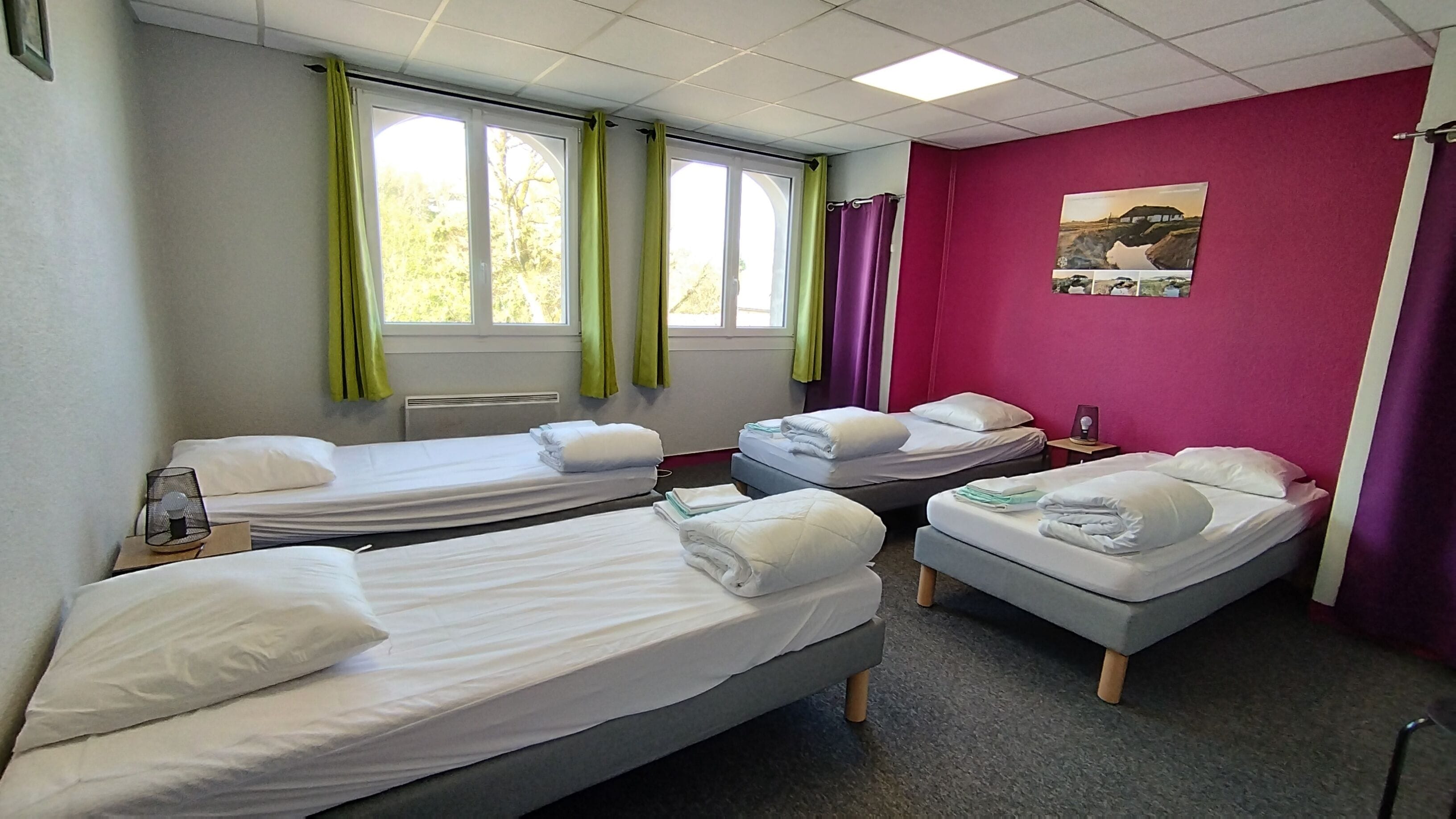 Ein Hotelzimmer mit drei Einzelbetten, einem fensterbelaubten Fenster, einem Heizkörper und einem Wandbild.