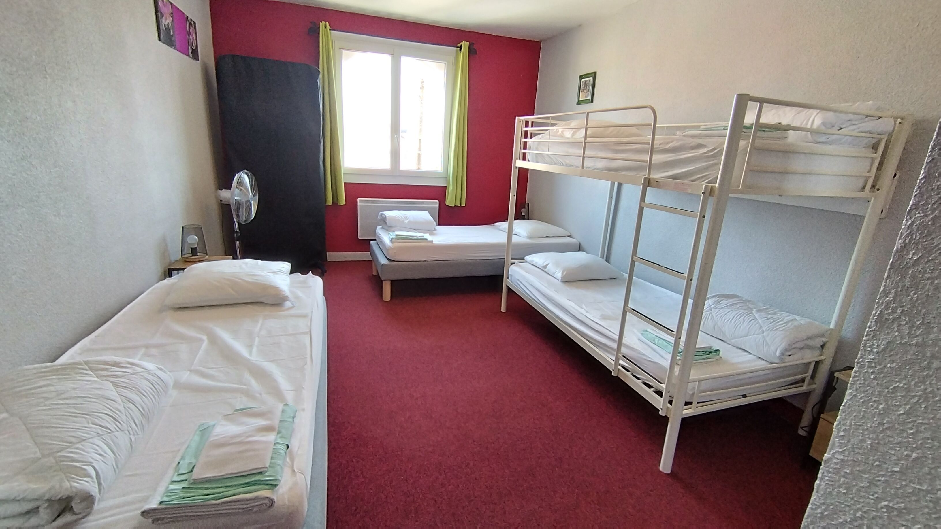 Une chambre avec des lits superposés, un lit simple, une fenêtre avec des rideaux, un ventilateur et une moquette rouge.