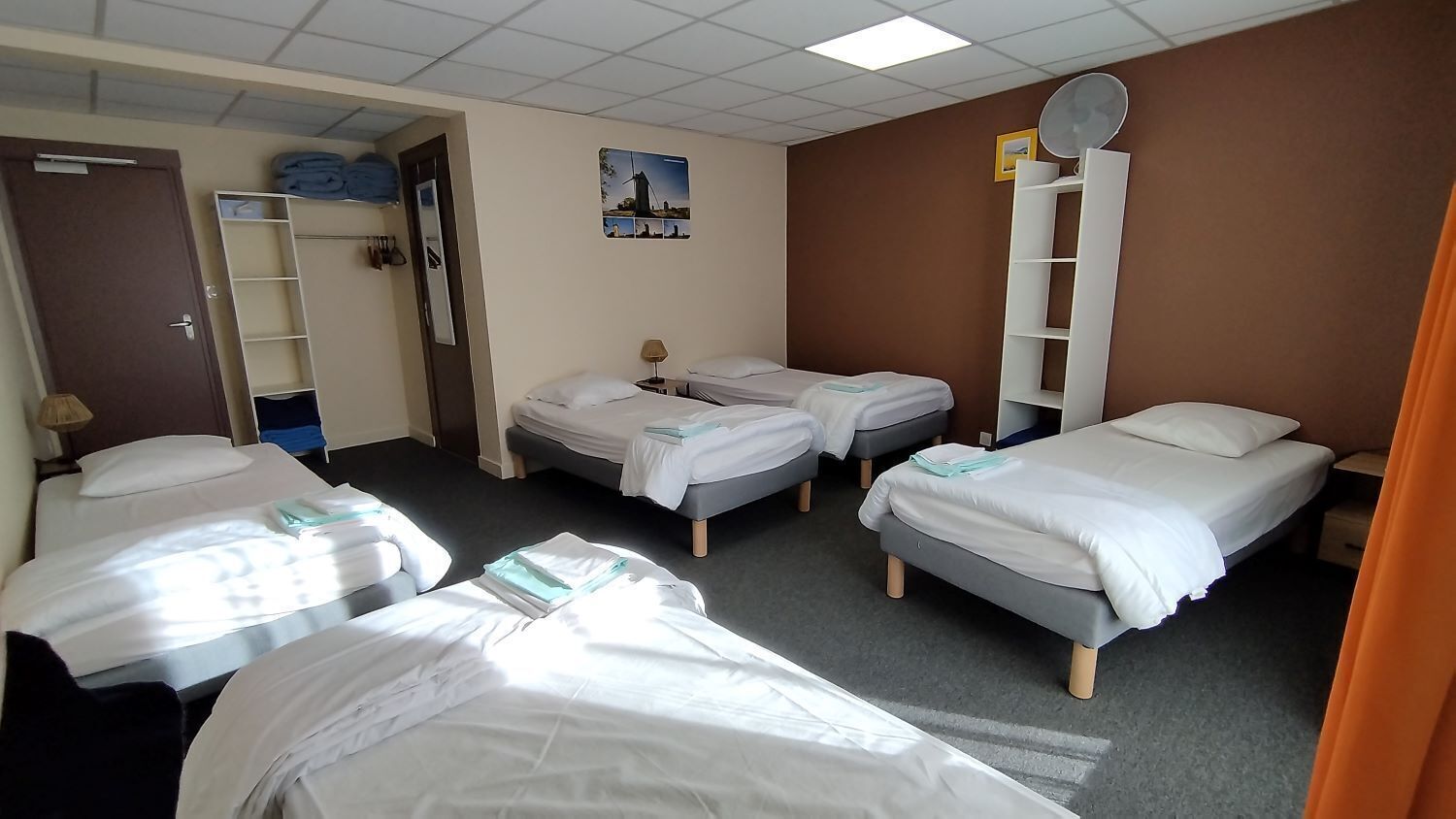 Une chambre d’hôtel avec quatre lits, une armoire, un miroir et un tableau représentant un moulin à vent accroché au mur.