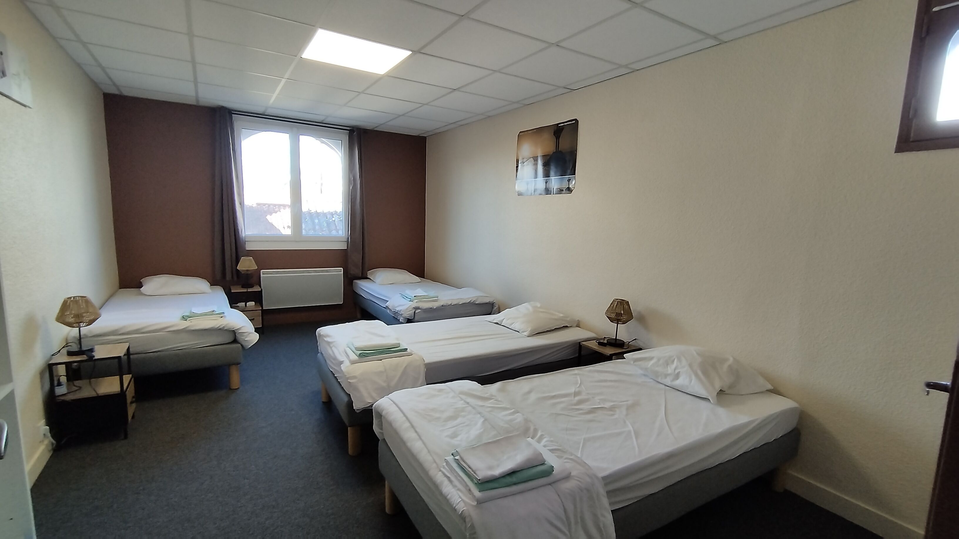 Habitación de hotel con dos camas individuales, una cama doble, una ventana con cortinas, un radiador y un cuadro pequeño en la pared.