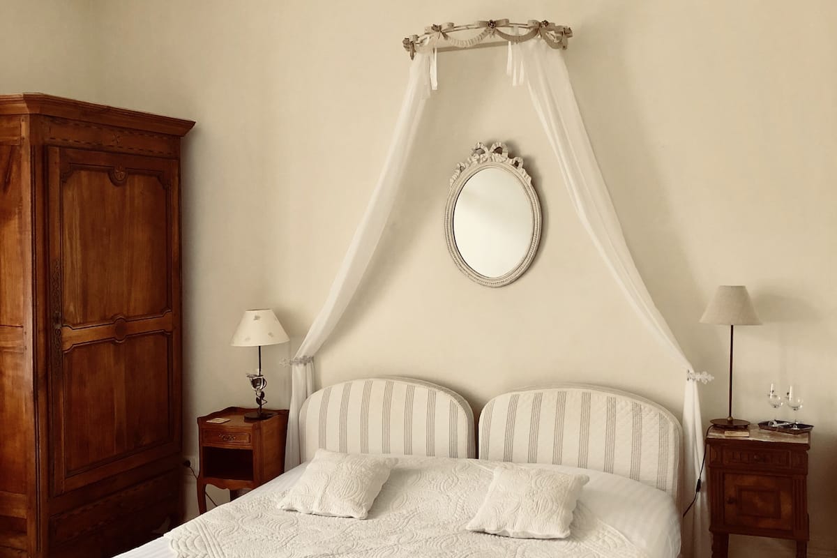 A bedroom with a four-poster bed, a wooden wardrobe, two bedside tables with lamps, and a mirror on the wall.