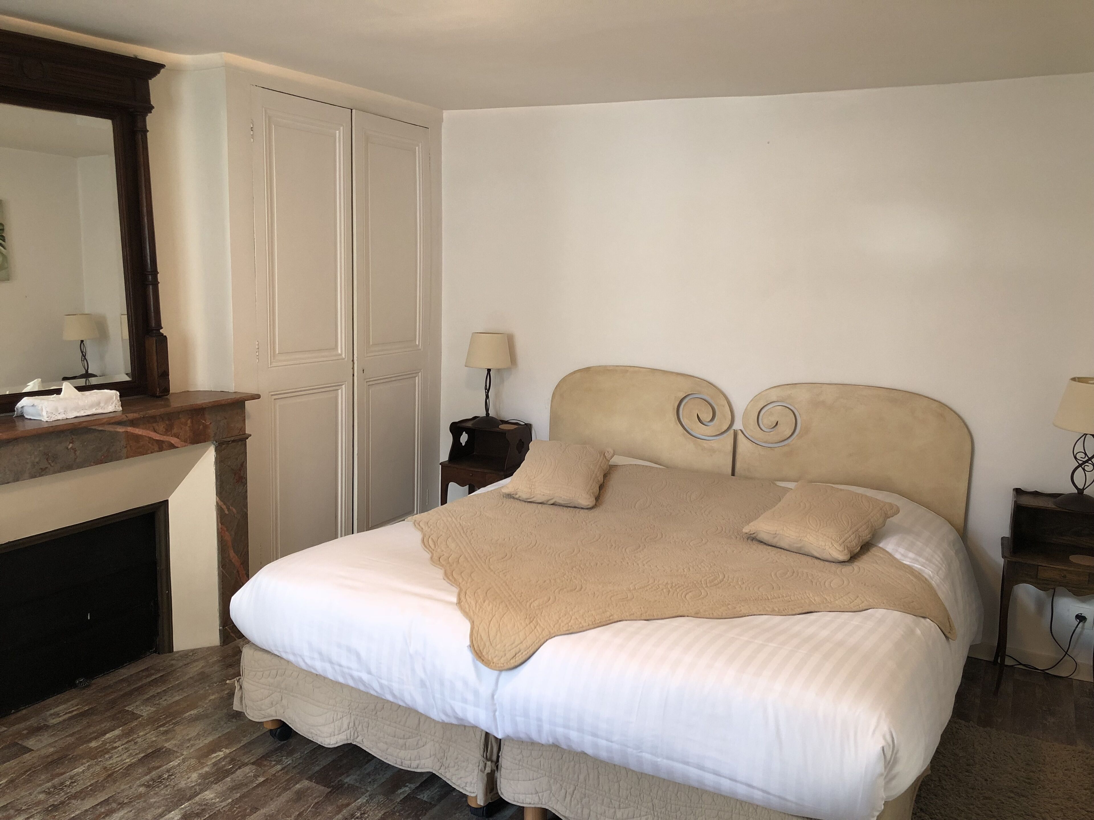 A neatly arranged bedroom with a bed, a nightstand, a lamp, a mirror, and a fireplace.
