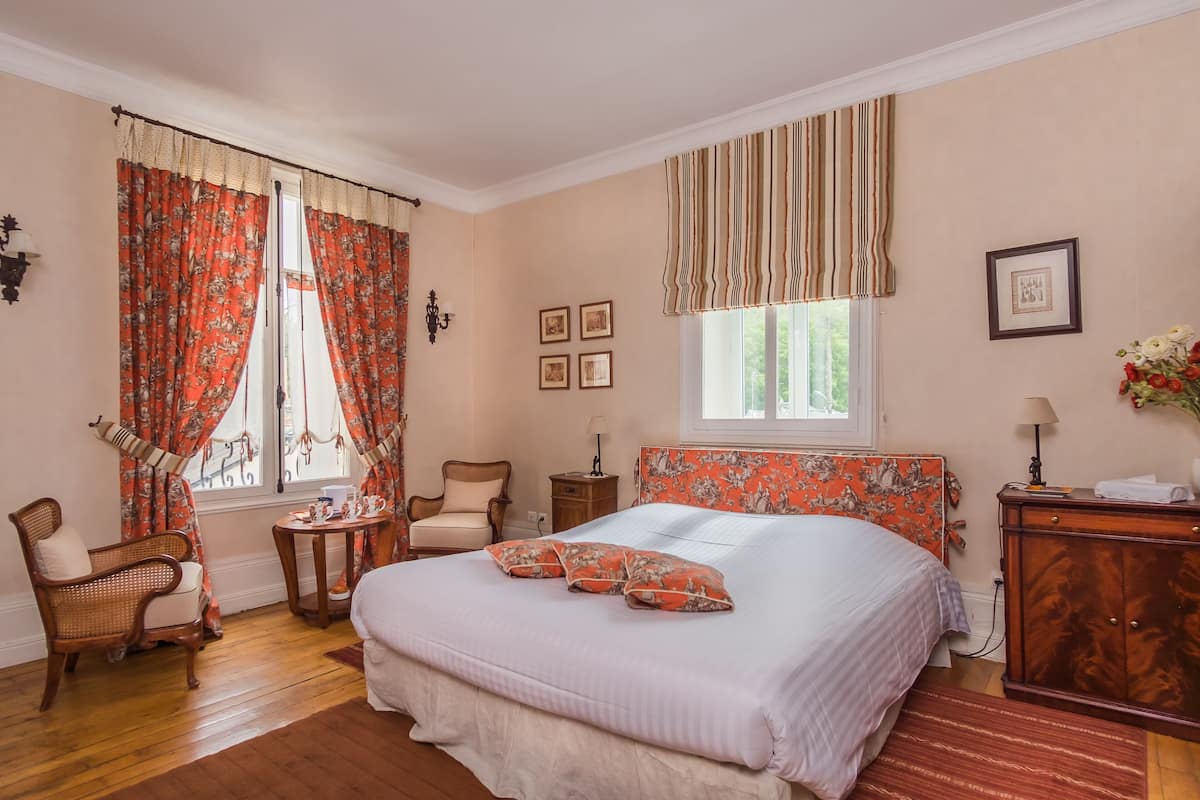 A bedroom with a large bed, two chairs, a small table, a nightstand, a dresser, and a window with curtains.