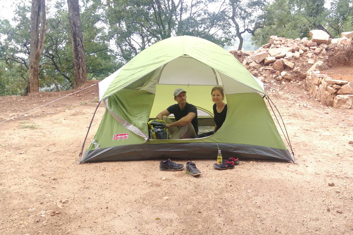 A green Coleman tent with two people inside, set up on a dirt ground with trees and a stone wall in the background.