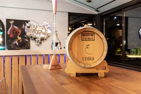 A wooden barrel labeled "BOURGOIN COGNAC L'ESSILLE" on a wooden stand in a bar setting with a modern interior and a glass display case in the background. - Logis Hôtel l'Essille (Bassac)