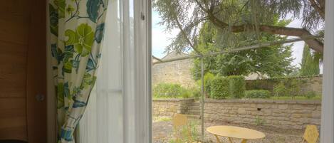 A room with a window, a wooden chair, a small round table, and a view of an outdoor garden with a stone wall and trees.