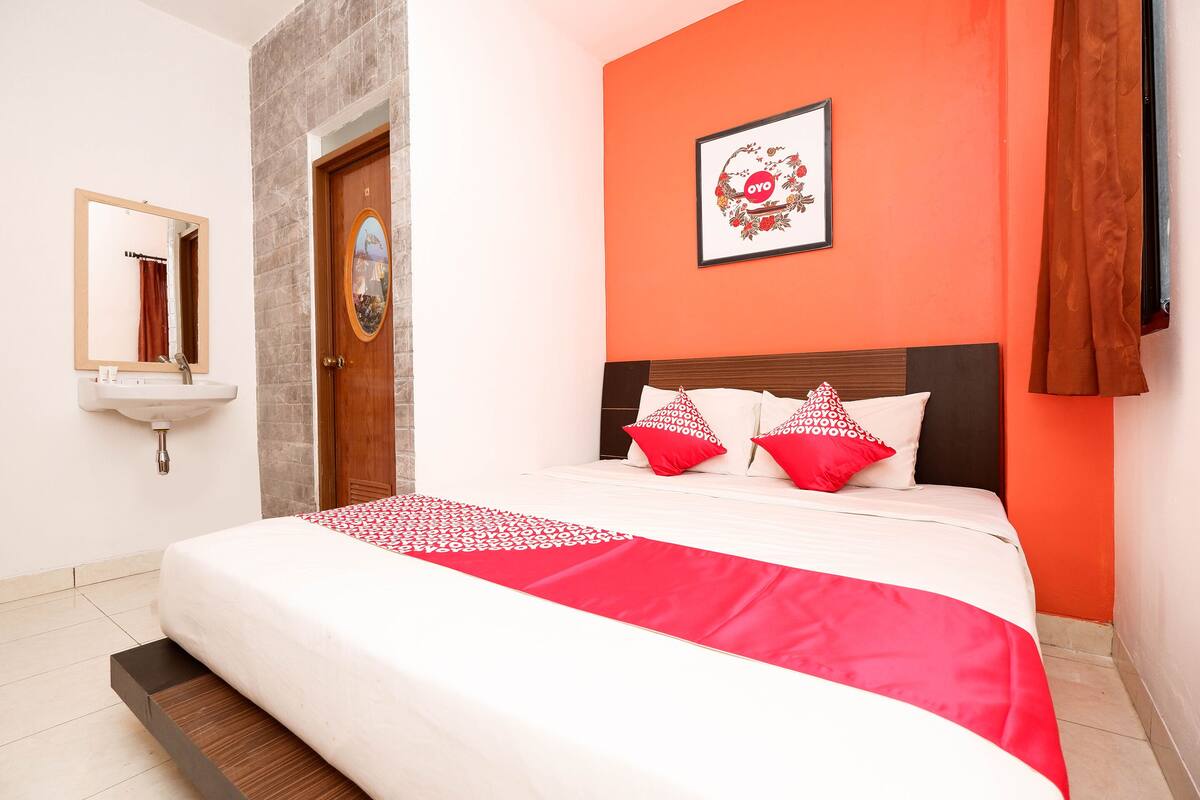 A hotel room with a bed, a wooden headboard, a red and white patterned bedspread, and a framed picture on the wall.